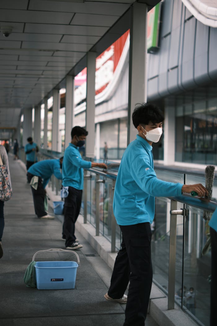 A team of workers in blue shirts with face masks clean glass railings outdoors during the day.